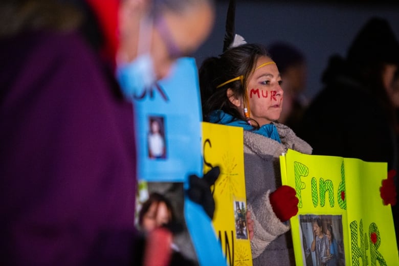 A woman wearing winter clothse stands among a crowd of others outsides, holding a sign reading "Find Her."