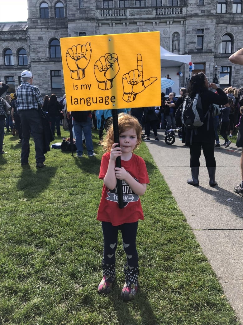 A girl in a red t-shirt and black leggings is pictured on a grass lawn, holding up a yellow sign that reads, "ASL is my sign language," with the letters A, S and L presented in illustrated sign language.