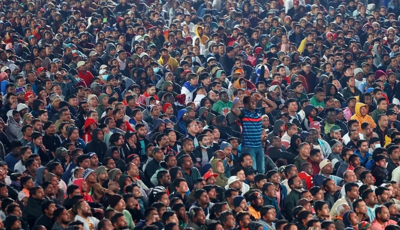 A smiling man stands with his arms raised and his hands close to his head while a large number of people are seated on the ground around him. They are all looking straight ahead in the same direction.