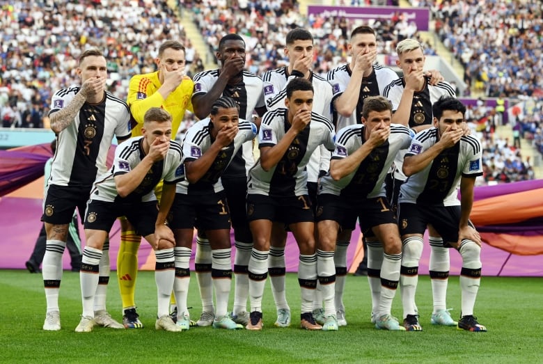 11 men in white and black uniforms lean forwards with their right hands over their mouths while standing close together on the grass of a soccer pitch.