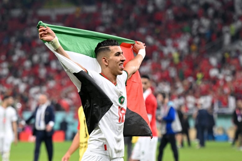 A smiling man in a white soccer shirt and shorts raises a green, white, black and red flag behind his back on a soccer pitch.