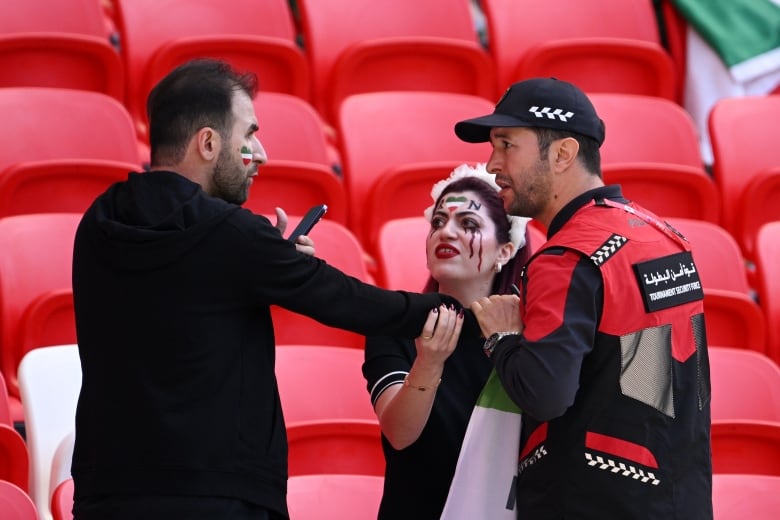 Three people - a woman and a man dressed in black with hearts painted on their faces in the colours of the Iranian flag, and a man wearing a jacket that says "tournament security police" - have a heated discussion in front of red chairs in the stands. The police officer appears to be trying to take a flag off the other man, while the woman grabs that man's arm.