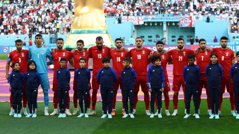 10 men in red T-shirts and shorts, and an 11th man wearing light blue, stand in a line with their arms around each other on the grass of a soccer pitch. Ten children dressed in dark blue sweatsuits stand in a line in front of the men.