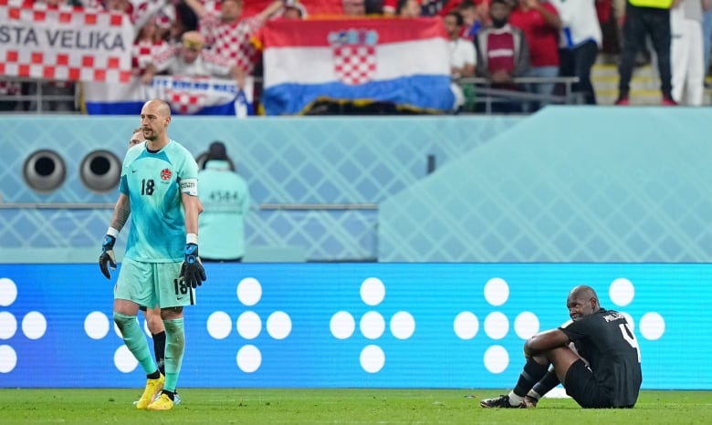 A man dressed in an aquamarine T-shirt and shorts, with gloves on his hands, walks on grass while another man dressed in black sits on the ground holding his knees. Red, white and blue flags are visible in the background.