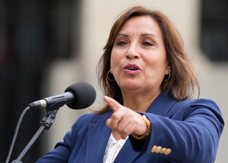 A woman in a blue blazer gestures while speaking at a microphone. 