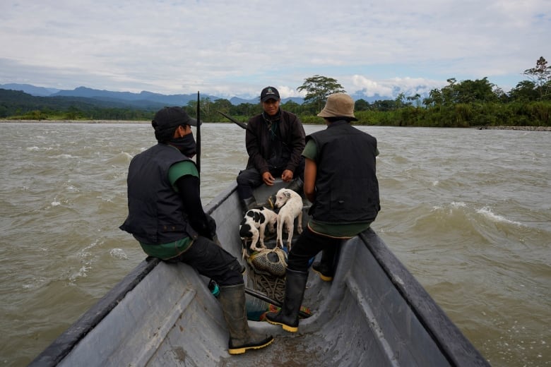 Three men sit on a small boat in the middle of a river, holding rifles. Two dogs sit at their feet.