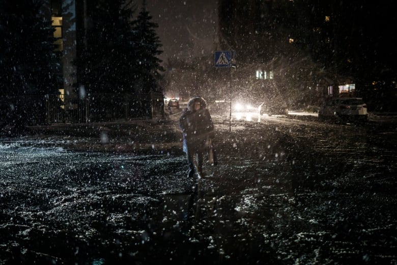 A figure in the middle distance, hood up, walks through the dark, with buildings, trees and a vehicle with illuminated headlights in the background.