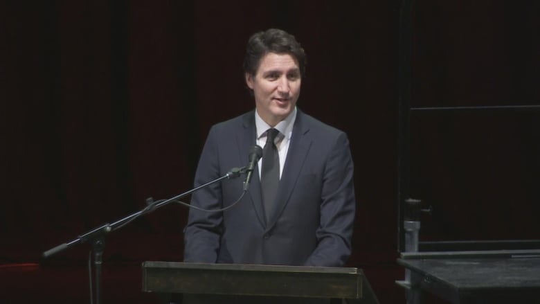 A man wearing a black suit and tie smiles slightly as he speaks at a podium.