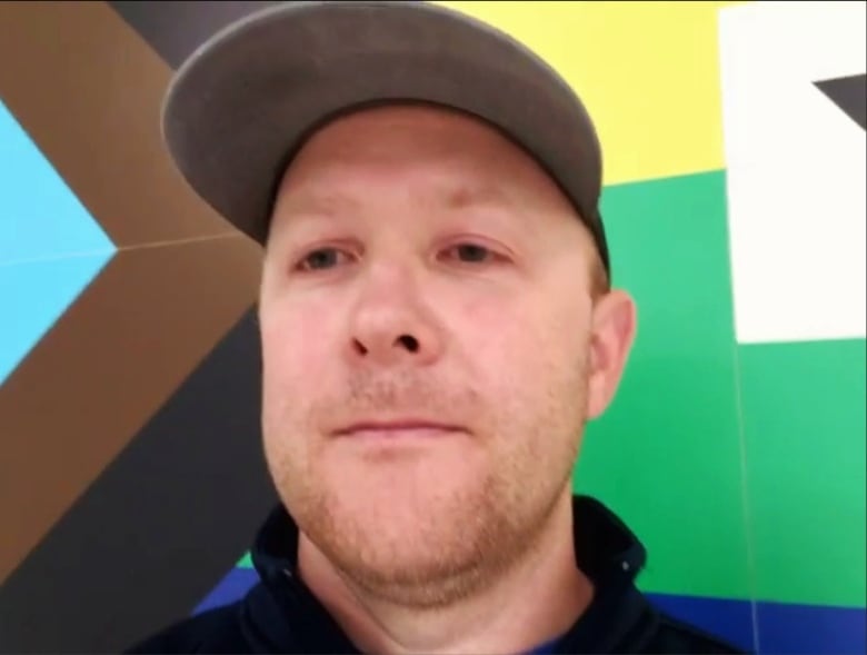 A man wearing a baseball cap stands in front of a mural of a pride flag.