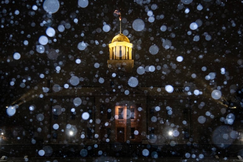 The building is mostly not visible in the dark, with only the top of the building and a flag brightly lit. The snowflakes are out of focus and appear as white circles.