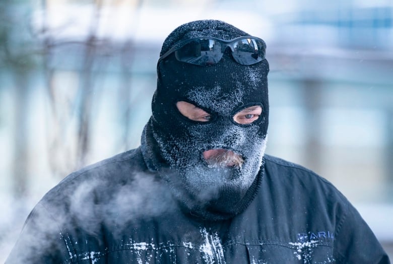 A man in a balaclava with sunglasses resting on the top of his head is pictured from the chest up. His frost-covered balaclava is slightly askew and his breath is visible.