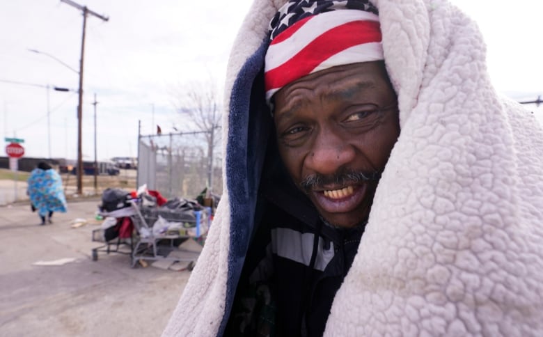 A man grimaces with a heavy blanket over the top of his head. A U.S. flag pattern on cloth on his head peeks out from under the blanket.