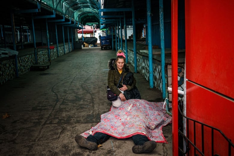 A woman kneels in front of a body that is covered by a blanket in a darkened passageway.