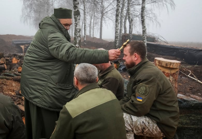 A man presses a gold cross to the head of a man in a green military jacket, sitting beside other men outside.