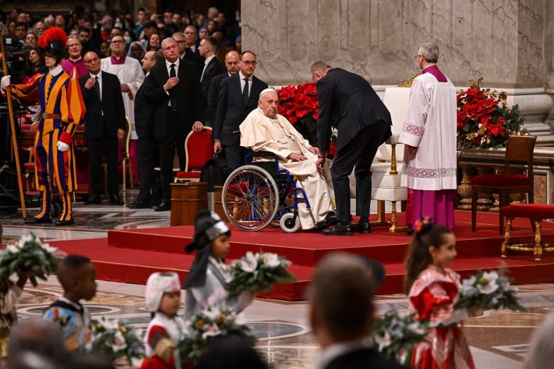 A man in a white robe sits in a wheelchair, surrounded by people.