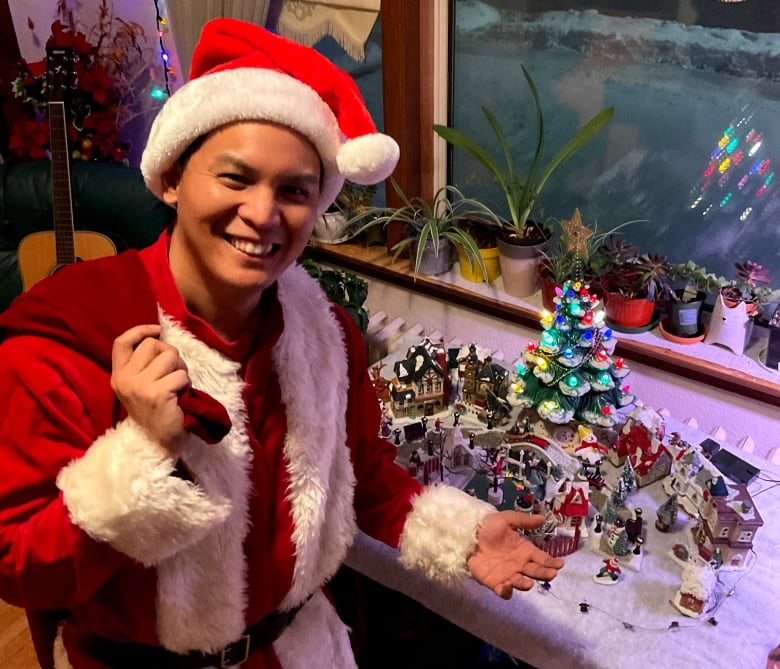 Jim Agapito, dressed as santa claus and with a sack slung over his shoulder, poses in front of a Christmas village display on a table.