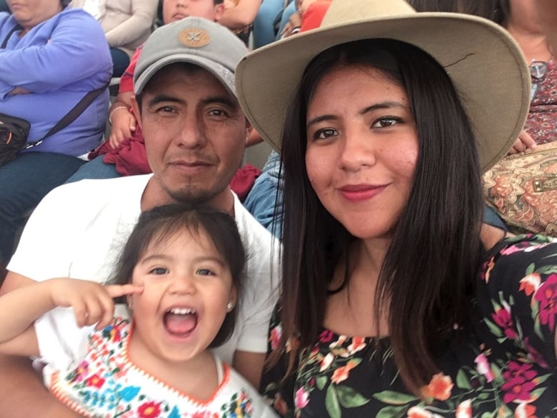 Three family members smile, posing for the camera at an event in Mexico. Salomon Lázaro holds his young granddaughter, who wears a colourful flowered Mexican blouse.