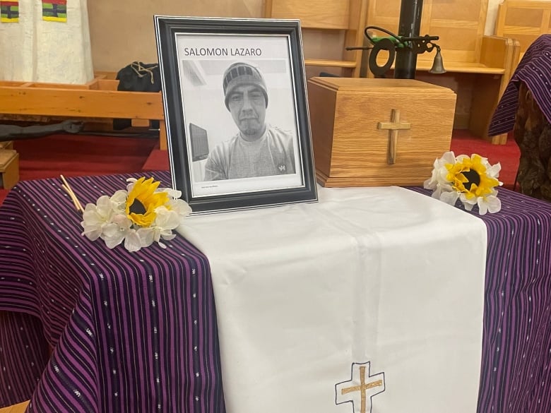 A black and white photo of Salomon Lázaro seen in the foreground. Beside it to the right is a wooden box with a cross containing his ashes. Black velvet bags can be seen in the background on the other side. They contain the ashes of other undocumented workers who also died of COVID-19