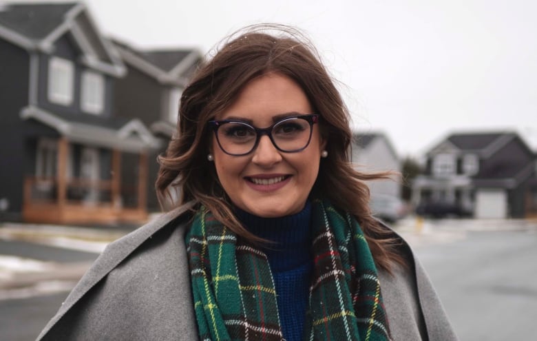 A woman wearing a grey coat and green scarf stands on a residential street.