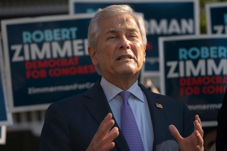 A man with grey hair, wearing a suit, speaks in front of a lot of signs saying 'Robert Zimmerman'.