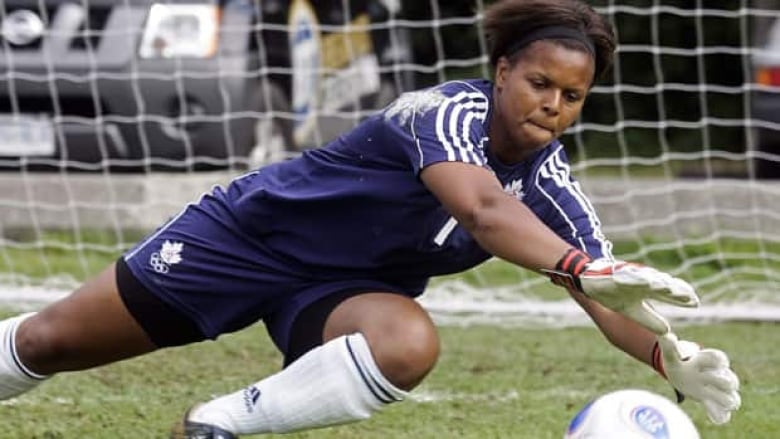 A woman in a soccer uniform dives to stop a ball from entering the goal. 