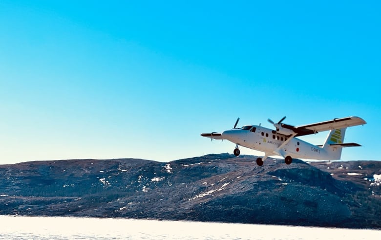 A plane flies over ocean ice framed against a deep blue sky and some rocky hills. 