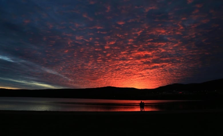 Two people can be seen close together in silhouette in front of a vibrant, yet dark, orange sunset, with the colors of the sky reflected off the ocean waters.