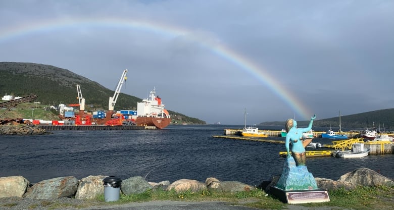 A statue of a mermaid in front of a harbor is framed so that a rainbow in the sky appears to be coming out of its hand.