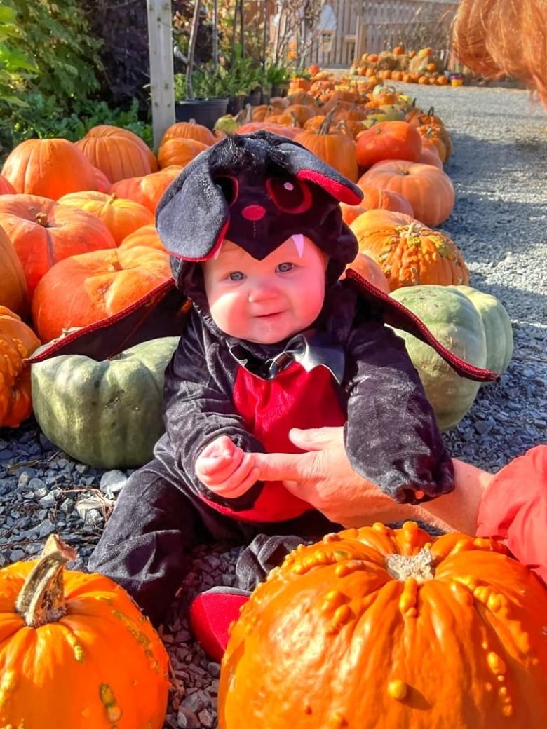 A baby in a bat costume, sitting in a pumpkin patch, smiles for the camera.