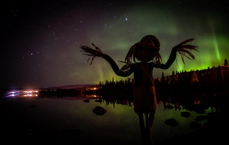 A scarecrow stands by the still waters in Labrador West, the northern lights in the sky above.