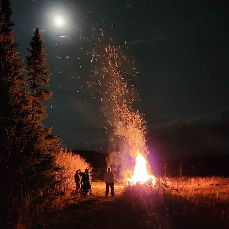 A plum of fire shoots up into the sky from a roaring bonfire, with a handful of people gatherer around at night.