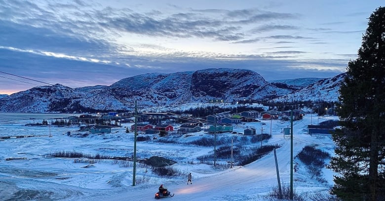 A wintery morning over a small community. Rooftops dusted with snow, mountain range in the background. Near the foreground, a person driving a skidoo passes a pedestrian. 