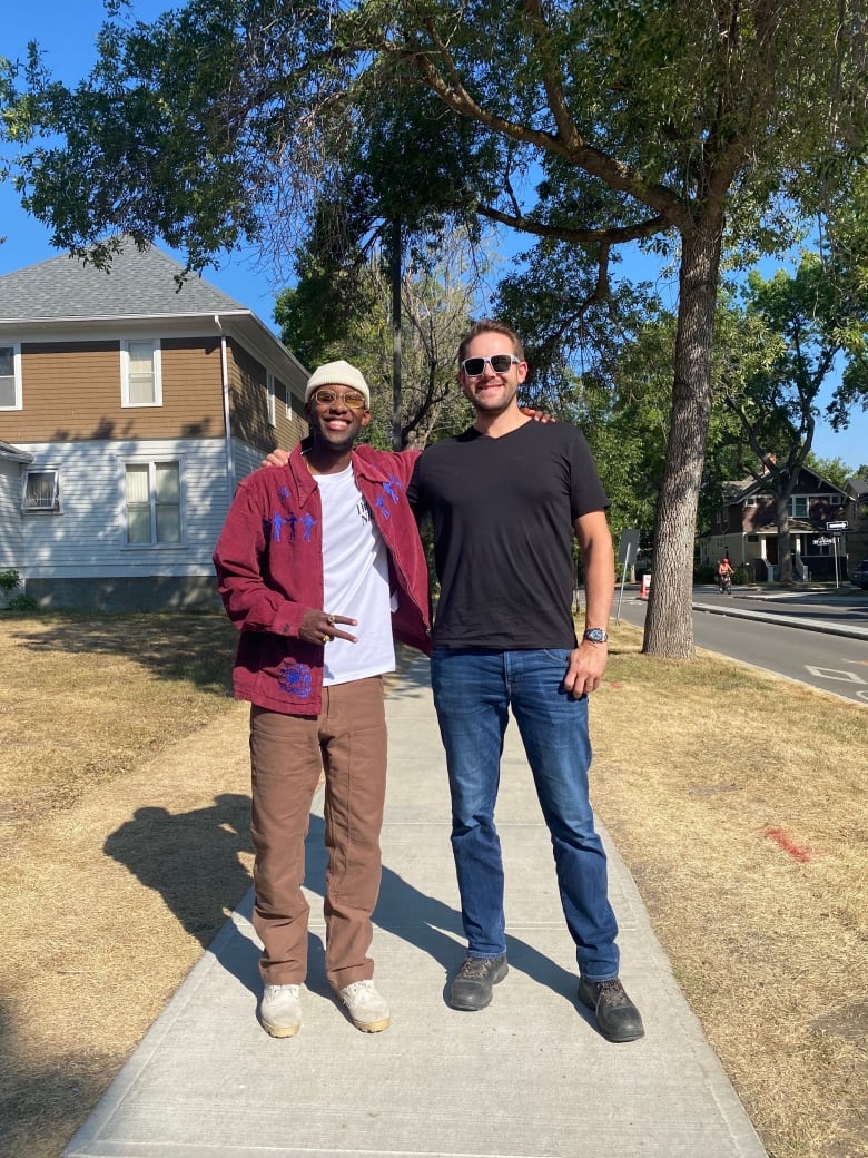 Two smiling men standing with arms around each other outside on a sunny day. 