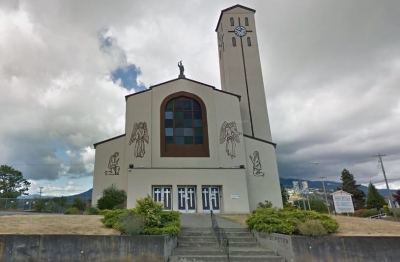 The light beige exterior of a Catholic church shows paintings of angels on the walls, crosses on the doors and a bell tower with a clock.