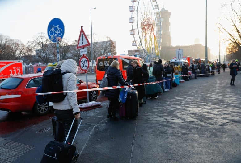 A long, cordoned line of people on the sidewalk beside a row of cars, a ferris wheel seen in the background.