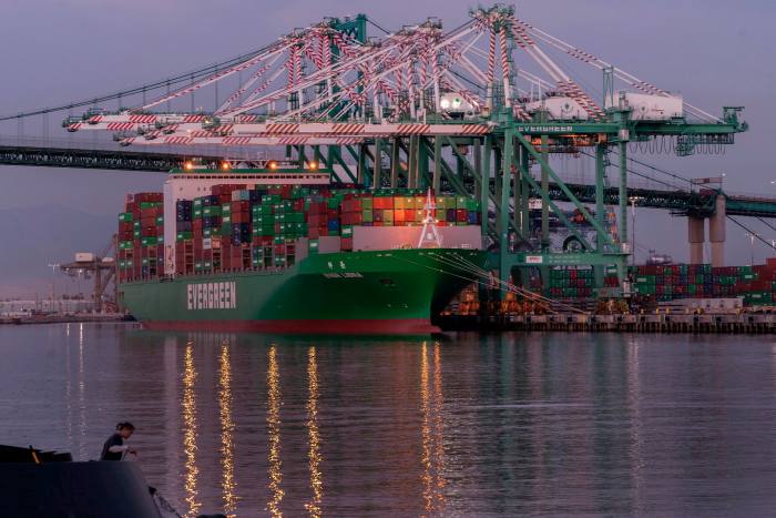 A large green container ship is moored at port, its orange lights reflected on the water