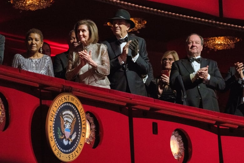 Several people are seen in box seats at a musical venue that is draped in red with a presidential seal on it. 