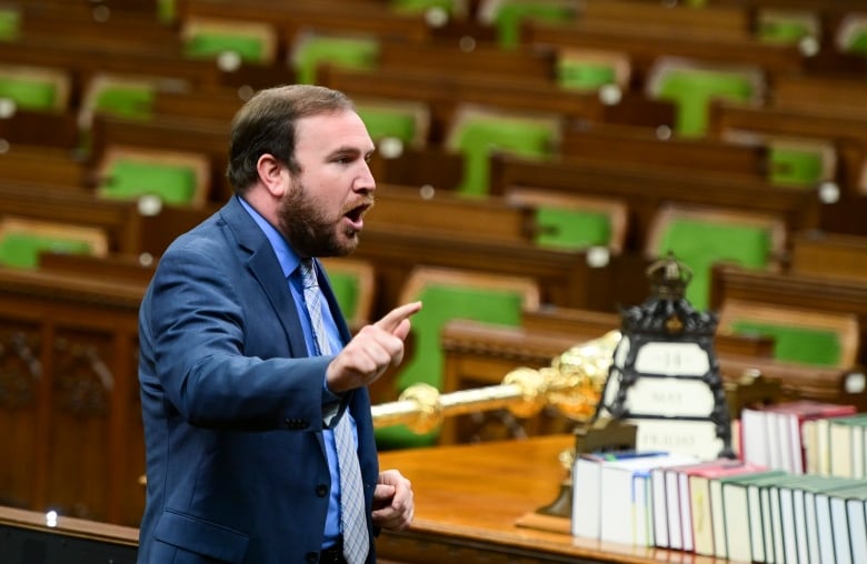 A bearded man in a blue suit speaks surrounded by empty seats.