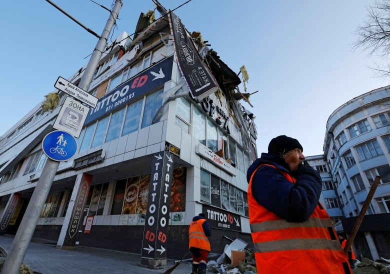 Workers in red vests work outside a damaged multi-storey building that houses a tattoo parlour at street level.