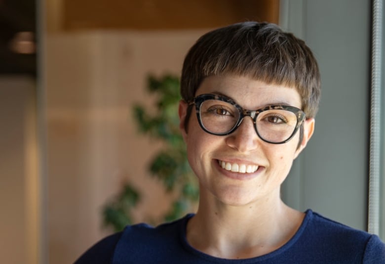 A woman, wearing a blue t-shirt and tortoise-shell cat-eye glasses, poses for a photo with her hand on her waist.