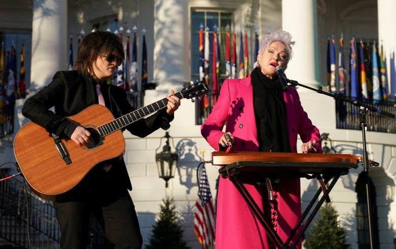 Individual with short, spiked pink hair wearing a hot pink coat, plays a keyboard and signs outside the White House. 