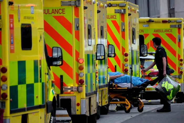 An ambulance worker wheels a patient on a trolley between queued emergency vehicles