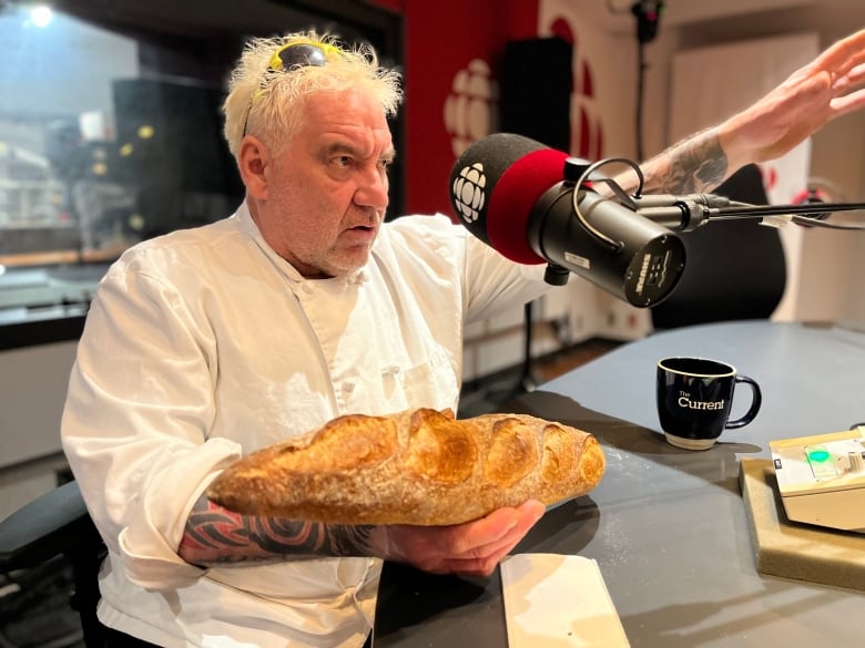 A white-haired man sits in a radio studio behind a microphone with the CBC logo on it. He's wearing a white chef's uniform, holding a baguette in one hand, and gesturing with the other. 