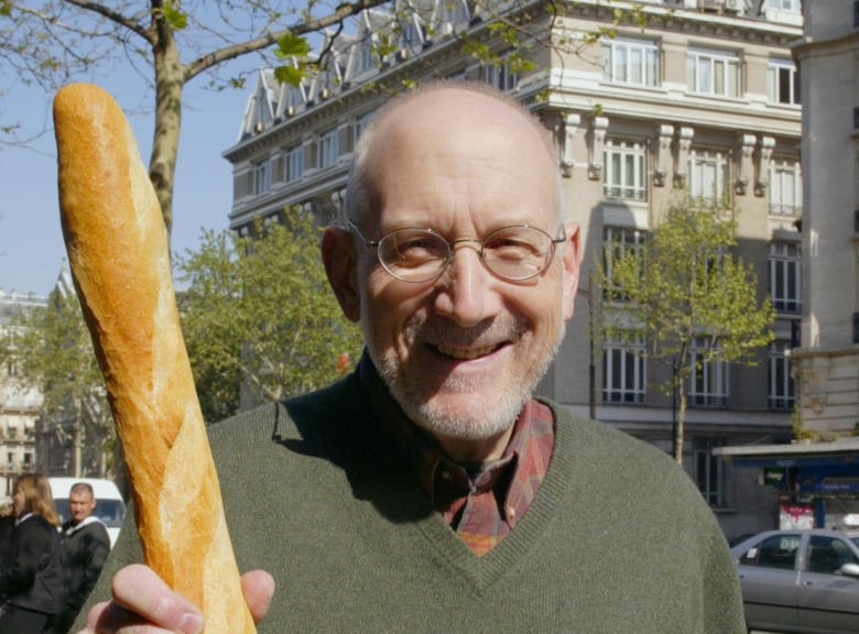 A smiling bald man with a gray beard and a green sweater stands on the streets of Paris holding a baguette in one hand and a book called Cherchez le Pain in the other.