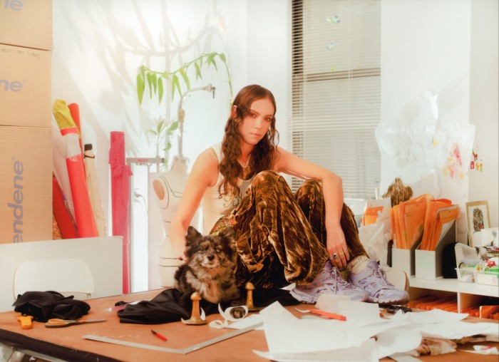 A young woman in a white T-shirt sits on large table with a small dog and is surrounded by the paraphernalia of a clothes designer — scissors, paper rulers