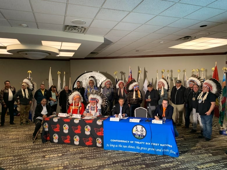 Four Treaty 6 chiefs, two wearing ceremonial headdresses, sit at a table while 16 other chiefs stand behind them in a hotel meeting room.