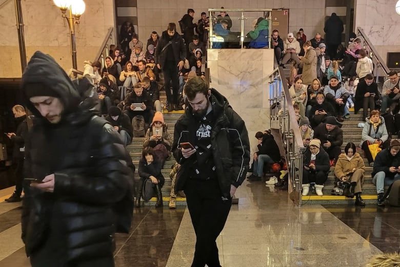 Dozens of people, many sitting on stairs, are shown in winter clothing inside a building.