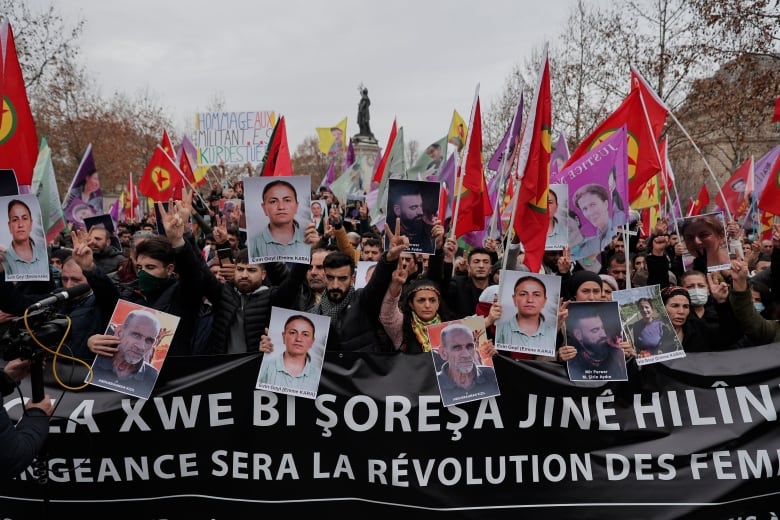 People wave flags and hold portraits and a long black banner with white lettering on a street in the daytime.