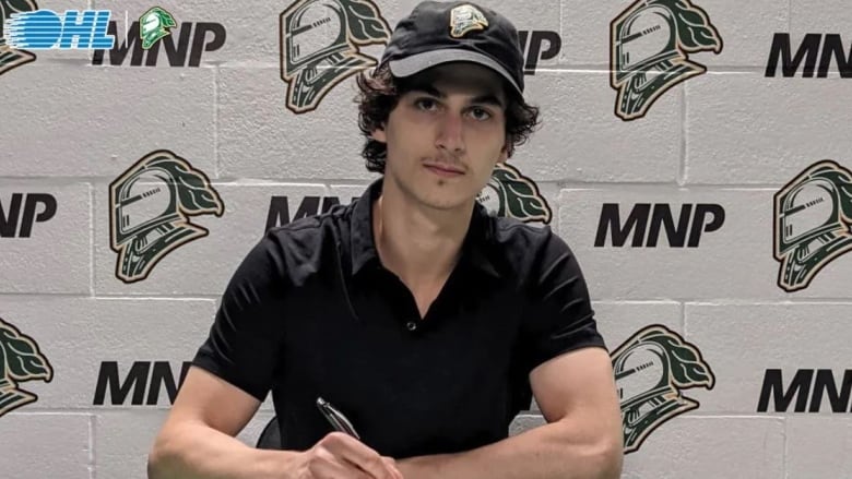 A young hockey player is seen signing a contract and looking at the camera while wearing a London Knights hat.