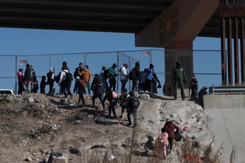 A group of people climb up a rocky to a fence where a line of people and border guards are standing.